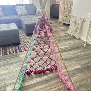 May include: A handmade, colorful teepee-style play tent. The tent features wooden poles and a macrame net in shades of pink, purple, and teal. White pom-poms and tassels add decorative accents. A fun and inviting space for children.