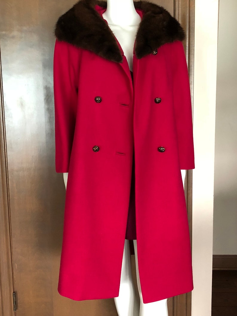 Puede incluir: Un abrigo largo de lana roja con un gran cuello de piel marr&oacute;n y cuatro botones decorativos.