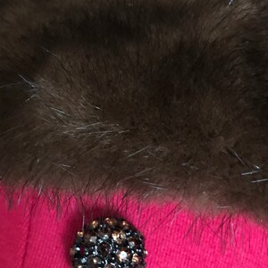 Puede incluir: Primer plano de un cuello de piel marr&oacute;n oscuro sobre una prenda rosa brillante. Un bot&oacute;n grande, redondo, negro con pedrer&iacute;a est&aacute; centrado en la prenda.