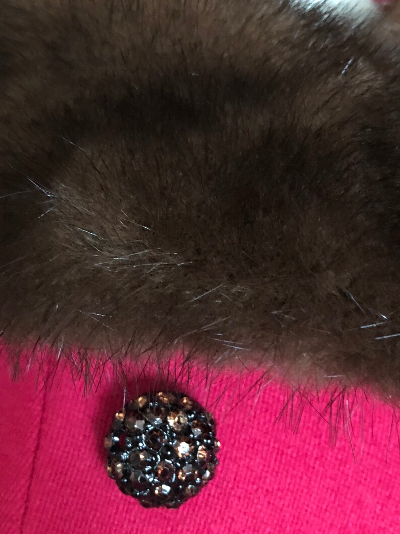 Puede incluir: Primer plano de un cuello de piel marr&oacute;n oscuro sobre una prenda rosa brillante. Un bot&oacute;n grande, redondo, negro con pedrer&iacute;a est&aacute; centrado en la prenda.