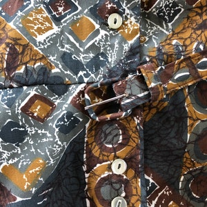 May include: Close-up of a patterned fabric with a geometric design in shades of brown, gray, and gold. The fabric features a repeating pattern of squares, diamonds, and circular shapes. White buttons and a belt with a silver buckle are visible.