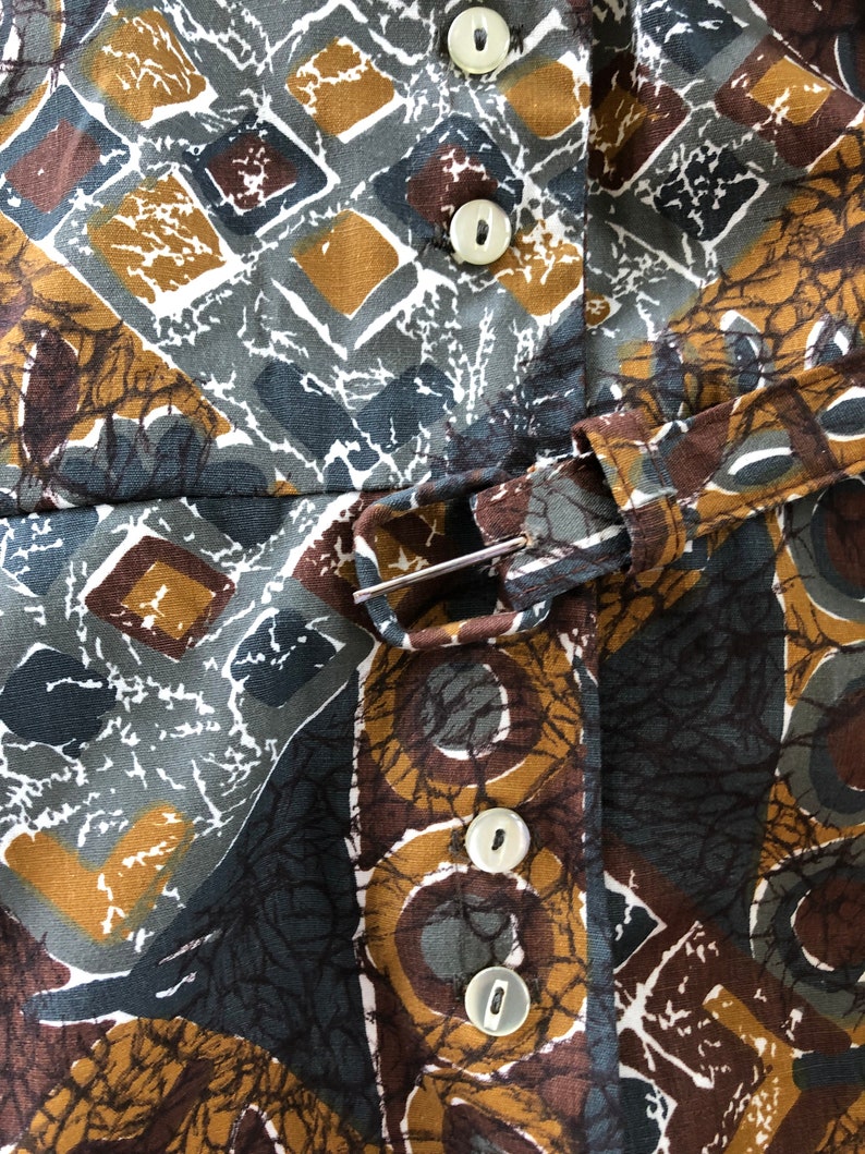 May include: Close-up of a patterned fabric with a geometric design in shades of brown, gray, and gold. The fabric features a repeating pattern of squares, diamonds, and circular shapes. White buttons and a belt with a silver buckle are visible.