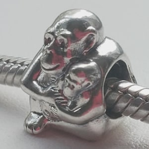 May include: A silver charm depicting a gorilla cradling a baby gorilla. The charm is detailed with facial features and fur texture. It is designed to be strung on a bracelet or necklace. The charm is made of a shiny, metallic material.