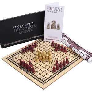 Puede incluir: Un tablero de juego de Hnefatafl de madera con piezas rojas y blancas. El tablero está rodeado por un borde decorativo. El juego está listo para jugar con las piezas en sus posiciones de inicio. El juego está empaquetado en una caja negra con el nombre "Hnefatafl" impreso en él. El juego también incluye un paño blanco con un patrón rojo y blanco.