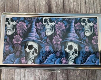 May include: A rectangular metal case featuring a repeating pattern of skulls wearing purple pointed hats, interspersed with purple and blue floral elements. The case has a polished silver-tone frame and a hinged lid with a glossy printed surface.