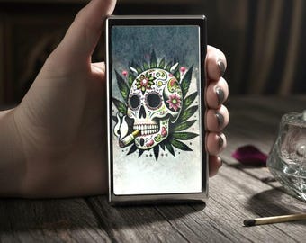 May include: A rectangular metal cigarette case featuring a colorful sugar skull graphic with floral details, surrounded by green cannabis leaves, and a smoking cigarette in the skull's mouth against a textured light gray background.