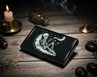 May include: A rectangular black metal cigarette case featuring a white graphic illustration of a skeleton sitting on a cloud, smoking, and surrounded by stars and a crescent moon. The case has a smooth, polished finish and a hinged lid design.