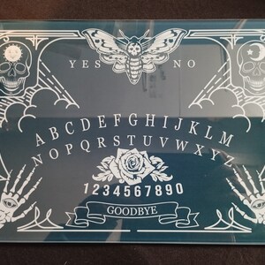 Puede incluir: Un tablero de Ouija azul verdoso con un diseño blanco de calavera, polilla y rosa. El tablero presenta el alfabeto, los números del 1 al 9 y el 0, y las palabras "YES", "NO" y "GOODBYE".