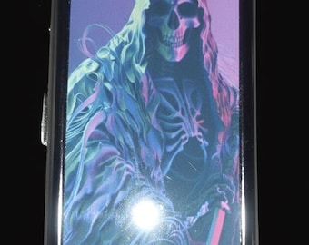 May include: A rectangular metal cigarette case featuring a vibrant graphic print of a hooded skeleton figure. The artwork displays a color palette of purple, blue, and pink hues. The case has a polished silver-toned frame and a smooth finish.