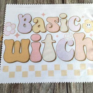 May include: A white square microfibre cleaning cloth with pinked edges featuring the text "basic witch" in large, rounded, pastel-coloured bubble letters. The design includes small flower icons and a chequered pattern along the bottom edge.