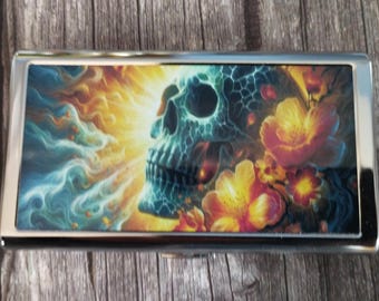 May include: A rectangular metal case featuring a vibrant graphic print on its lid. The design displays a blue and teal skull surrounded by bright yellow and orange flowers, set against a background of swirling light rays and smoke-like patterns.