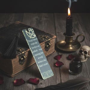 Puede incluir: Imagen de temática gótica con un marcapáginas con la frase: «El mayor truco que hizo el Diablo fue hacer creer al mundo que no existía». Vela negra, calavera, tintero y pluma.