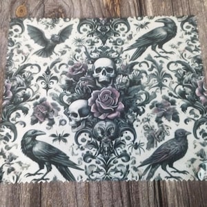 May include: A square microfibre cleaning cloth featuring a repeating gothic pattern with black ravens, a bat, skulls, and muted purple roses set against a white background. The edges of the cloth are finished with a decorative pinked zigzag cut.