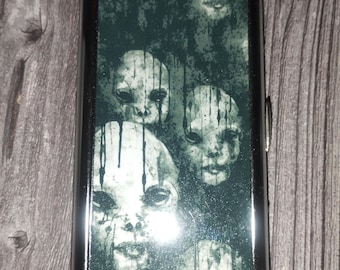 May include: A rectangular metal cigarette case with a polished dark frame. The front panel features a dark, monochromatic graphic print depicting multiple pale, stylized faces with dripping ink effects, creating a layered and haunting visual composition.