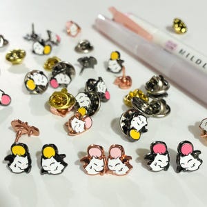 May include: A collection of enamel pins featuring a cartoon cat design. The pins are in various colors, including black, white, yellow, pink, and gold. Some pins have a butterfly clasp. Pens are visible in the background.