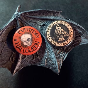 May include: Two round button pins. One pin is orange with black text that reads "Everyday is Halloween". The other pin is black with gold text that reads "Proud Member Weirdo Society".