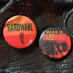 May include: Two round button pins with the word "HARDWARE" printed on them. The first pin has a red background with a silhouette of a person walking in a desert landscape. The second pin has a black background with a skull and the text "M.A.R.K. 13" above the word "HARDWARE".