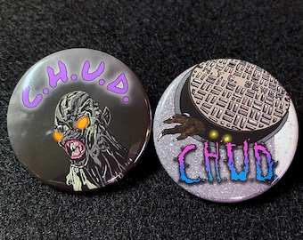 May include: Two round buttons with a black background and a white design. The first button features a creature with glowing orange eyes and the text "C.H.U.D." in purple. The second button features a sewer grate with the text "CHUD" in blue and pink.