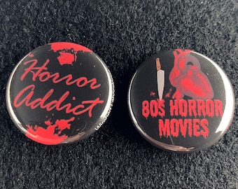 Puede incluir: Dos botones redondos negros y plateados con detalles rojos. El primer botón tiene el texto "Horror Addict" en blanco con salpicaduras rojas. El segundo botón tiene un corazón rojo con un cuchillo atravesándolo y el texto "80s Horror Movies" en blanco.