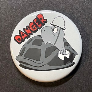 May include: A white button with a cartoon turtle wearing a hard hat and a bow tie. The turtle is in a shell and the button says "DANGER" in red letters.