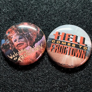 May include: Two round buttons with a horror movie theme. The first button features a close-up of a creature with the text "Welcome to Frogtown". The second button has a white background with the text "Hell Comes to Frogtown".