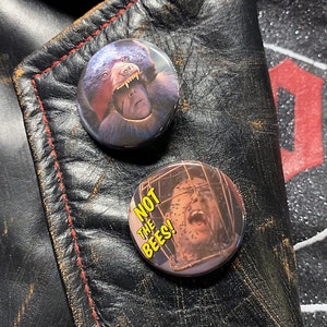 May include: Two round button pins with images from the movie The Texas Chainsaw Massacre. The first pin features a close-up of Leatherface's face with the text "Not the Bees!" in yellow. The second pin features a close-up of a man's face with bees swarming around him.