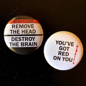 May include: Two round buttons with black and white text on a black background. The first button says "NEWS FLASH REMOVE THE HEAD DESTROY THE BRAIN". The second button says "YOU'VE GOT RED ON YOU" with a red splatter on the right side.