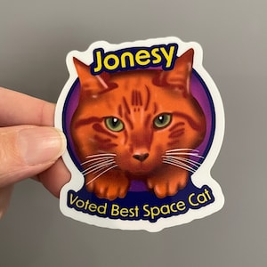 May include: A sticker featuring a cartoon orange tabby cat with the name "Jonesy" and the text "Voted Best Space Cat".