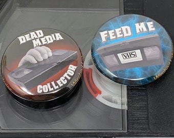 May include: Two circular pin badges featuring VHS tape designs. One badge says "DEAD MEDIA COLLECTOR" with a hand reaching for a VHS tape. The other badge says "FEED ME" with a VHS tape graphic and blue lightning bolts.