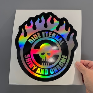 May include: A holographic sticker with a black border and the words "RIDE ETERNAL SHINY AND CHROME" encircling a skull and crosshairs design. The sticker has a flame-like border and rainbow color effects.