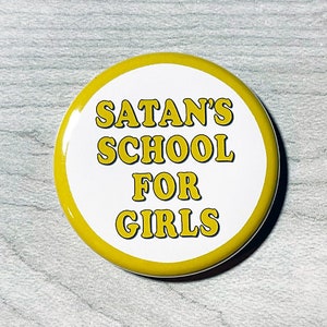 May include: A yellow button with the text "SATAN'S SCHOOL FOR GIRLS" in white letters.