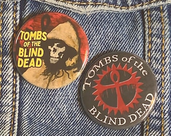 May include: Three button pins on a blue denim jacket. The pins are round and feature different designs. One pin is black with a red and white design and the text "Tombs of the Blind Dead". Another pin is black with a red and white design and the text "Wired Laboratories". The third pin is yellow with a black and orange design and the text "Tombs of the Blind Dead".