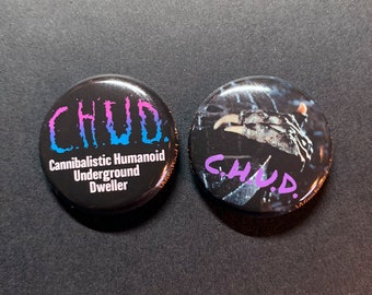 May include: Two round black button badges with a white background. The left badge has the text "CHUD" in pink and blue lettering. Below the text is "Cannibalistic Humanoid Underground Dweller". The right badge has a black and white image of a creature with the text "CHUD" in purple lettering.