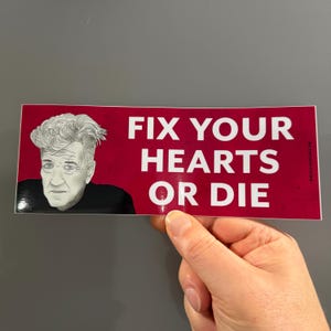 May include: A rectangular sticker with a red background and white text that reads "FIX YOUR HEARTS OR DIE." The sticker features a portrait of a man with white hair and a black shirt.
