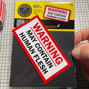 May include: A black and white sticker pack with a red and white warning label that reads "WARNING MAY CONTAIN HUMAN FLESH". The sticker pack also includes a yellow backing with a logo and text.