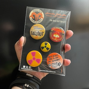 May include: A pin-back button set with a post-apocalyptic theme. The set includes buttons with text like "FALLOUT" and "THEY BLAST THE FLESH OFF HUMANS!" in a variety of colors, including yellow, orange, and black. The buttons are packaged on a black backing.