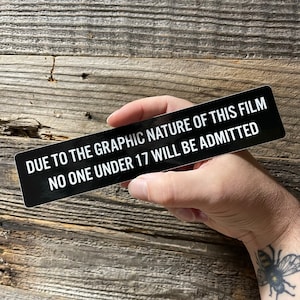 May include: Black and white sign with the text "DUE TO THE GRAPHIC NATURE OF THIS FILM NO ONE UNDER 17 WILL BE ADMITTED".