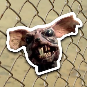 May include: A close-up of a snarling, grotesque creature with large ears and sharp teeth. The creature is peeking through a chain-link fence.
