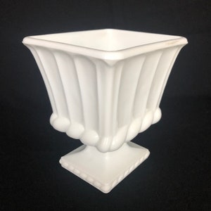 Puede incluir: Un jarrón de cerámica blanco con una parte superior cuadrada y un diseño acanalado. El jarrón tiene una base de pedestal con una parte superior cuadrada y un diseño de cuerda alrededor de la parte inferior.