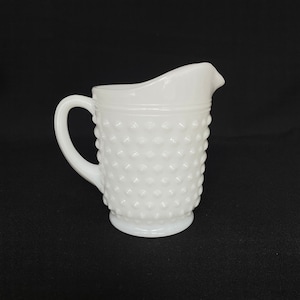 Könnte beinhalten: Weißer Milchglas-Krug mit einem strukturierten, erhabenen Punktmuster. Der Krug hat einen Henkel und einen abgerundeten Ausguss.