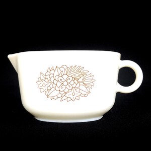 May include: Cream-colored vintage creamer with a brown floral design. The creamer has a spout for pouring and a handle for easy use. The floral pattern includes flowers and leaves. The creamer is made of ceramic.