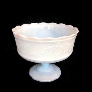 Puede incluir: Un cuenco de pedestal de vidrio blanco opalino con borde festoneado. El cuenco presenta un patrón detallado de puntos y diseños florales. La base con pie también está decorada con un patrón similar. El cuenco está sobre un fondo negro.