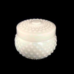 May include: A white hobnail jar with a matching lid. The jar and lid are a creamy white colour. The jar has a rounded shape with a textured surface of small, raised bumps. The lid is flat with a central knob and a similar hobnail pattern.