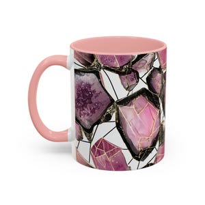 May include: A white ceramic mug with a pink handle and interior. The mug features a repeating pattern of purple and pink gemstone designs with gold accents and black outlines. The gemstones are set against a white background.