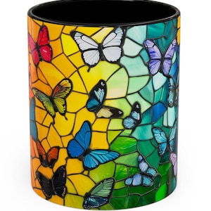 May include: A ceramic mug with a colorful stained glass design featuring butterflies in shades of blue, green, yellow, red, and purple. The mug has a black rim and handle.