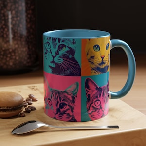 May include: A ceramic mug with a blue interior and handle, featuring a pop art design of four cat faces in teal, yellow, pink, and orange squares. The mug sits on a wooden surface with a spoon and a pastry.