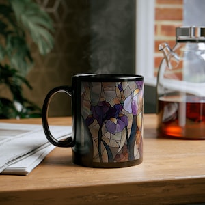May include: Black ceramic mug with a stained-glass effect design of purple and white irises. The mug has a curved handle and contains a hot drink, with steam rising. The mug is on a wooden surface, with a glass teapot in the background.