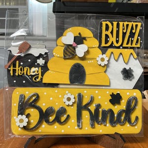 May include: A set of wooden decorative signs featuring a yellow beehive, a black honey pot with a dipper, and a yellow rectangular sign with white polka dots and the text Bee Kind. The set includes white flower accents and the word BUZZ.