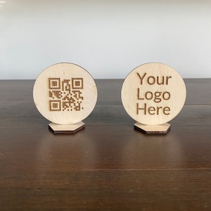 May include: Two wooden signs with rounded edges and hexagonal bases. The sign on the left has a black and white QR code. The sign on the right says "Your Logo Here".