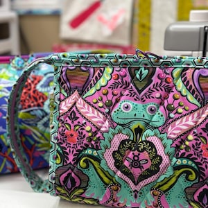 May include: A quilted fabric bag featuring a vibrant, repeating pattern of green frogs, pink floral motifs, and black geometric accents. The bag has a structured rectangular shape with a top zipper closure and a matching patterned shoulder strap.